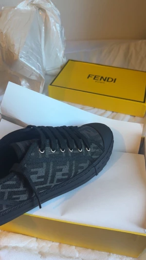 Svarta Fendi sneakers med mönster - Snygga svarta sneakers från Fendi med deras ikoniska mönster i grått på sidan. Skon har snörning med metallöglor och en platt sula i svart gummi. Materialet är textil med detaljer i syntet och skinn. Perfekt för dig som gillar exklusiv streetstyle.