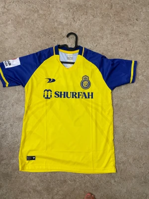 Al Nassr Ronaldo #7 fotbollströja - Säljer en officiell Al Nassr fotbollströja med Ronaldo och nummer 7 på ryggen. Tröjan är gul med blå detaljer och korta ärmar, tillverkad i lätt och ventilerande polyester. Klubbmärket och sponsorlogga finns på bröstet, samt tryck på ärmen.