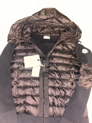 Svart Moncler pufferjacka med huva - Svart pufferjacka från Moncler med stickade ärmar och huva. Helt oanvänd och allt är bra nfc scanen har inga problem heller. Jackan har quiltad front, dragkedja och Moncler-logga på vänster ärm. Snyggt ribbade muddar och två fickor med knappstängning. Perfekt för dig som gillar exklusiva streetwear-vibbar. Skriv gärna om du har fler frågor mvh.