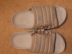 Beige Nike sandaler med snörning - Unika beige sandaler från Nike med vadderad ovandel och snörningsdetalj framtill. Skorna har öppen tå och platt, räfflad sula för extra grepp. Perfekta för sommaren och chill dagar. Diskret Nike-logga på ovandelen.