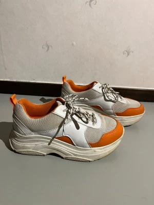 Chunky vita och orange sneakers - Säljer ett par chunky sneakers i vitt och orange med plattformsula. Skorna har meshpaneler för extra ventilation, detaljer i syntet och coola snören med svarta detaljer. Insidan och dragflikarna är i stark orange färg som ger en snygg kontrast. Knappt använda och i mycket bra skick!