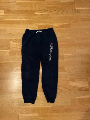 Mörkblå mjukisbyxor från Champion - Snygga mörkblå mjukisbyxor från Champion med logga och text längs benet. Byxorna har snörning i midjan och muddar vid bensluten för en skön passform. Perfekta för chill eller träning, tillverkade i mjukt bomullsmaterial.