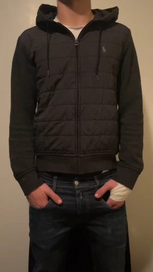 Svart pufferjacka från Polo Ralph Lauren - Svart pufferjacka från Polo Ralph Lauren med huva och snörning. Jackan har quiltad framsida, dragkedja och diskret logga på bröstet. Ärmar och rygg är i mjuk bomull, medan framsidan är i syntetmaterial. Perfekt för höst och vinter.