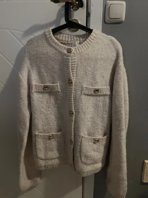 Beige stickad kofta med knappar - Mysig beige stickad kofta med framfickor, alla med dekorativa knappar. Koftan har långa ärmar och ribbade detaljer vid hals, fickor och nederkant. 