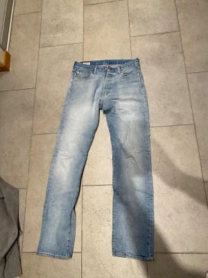 Levi's 501 ljusblå jeans W30 L34 - Klassiska Levi's 501 jeans i ljusblå tvätt med raka ben och femficksmodell. Jeansen har knappgylf och den ikoniska läderpatchen bak i midjan. Perfekt för dig som gillar en tidlös och avslappnad stil.