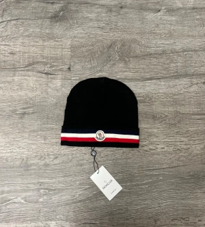 Mörkblå Moncler mössa med ränder - Snygg mörkblå mössa från Moncler med klassiska ränder i blått, vitt och rött längs uppviket. Mössan har Moncler-logga framtill och är stickad i mjukt material. Perfekt för dig som vill ha en stilren och sportig look.