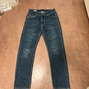True Religion Jeans -  säljer ett par blå True Religion jeans med ikoniska kontrastsömmar och stora bakfickor med lock och tryckknappar.Klassisk femficksmodell, tillverkad i USA av bomull Snygg tvätt och detaljer som sticker ut. De är även knappt använda och ett nytt par skulle kosta 2000-2500 så det är ett bra pris jag har satt men du kan även pruta