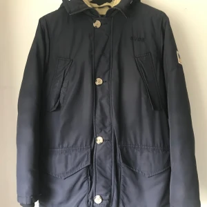 Vinterjacka Mörkblå från Svea  - Vinterjacka med mörkblått från Svea, stora fickor och knäppning med stora knappar.samt dragkedja. Jackan har hög krage och klassisk parkasdesign, perfekt för kyliga dagar.Det saknas fuskpäls på jackans huva.Annars hel och fin skick.