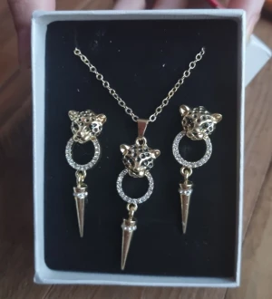 Smyckesset med leopardmotiv - Ett justerbart halsband och matchande örhängen. Smyckena har en design med leopardhuvuden och detaljer med strass och spetsiga hängen. Passar bra som present. Färg : gyllene Material : zink legering  Örhängens längd 4cm Asken ingår