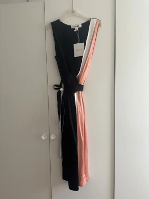 Svart omlottklänning från Diane von Furstenberg - Snygg omlottklänning från Diane von Furstenberg i svart, puderrosa och vit. Klänningen är ärmlös, har omlottdesign med knyt i midjan och är knälång. Materialet är mjukt och har en lyxig känsla, perfekt för dig som gillar stilrena och coola färgblock. Klänningen är i storlek US 8