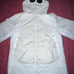 Vit pufferjacka från C.P. Company - Säljer en snygg vit pufferjacka från C.P. Company i storlek M. Jackan har en cool design med huva och stora fickor framtill. Den är gjord av polyester och har en dragkedja. Perfekt för vintern och håller dig varm och stilren! ❄️ 
