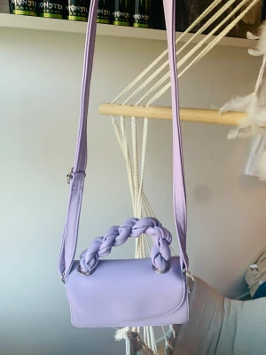 Ljuslila axelväska med flätat handtag - Superfin ljuslila axelväska i syntetmaterial med justerbar rem och ett snyggt flätat handtag. Väskan har ett stilrent och modernt utseende, perfekt för dig som vill sticka ut med en trendig accessoar. Rymmer det viktigaste och är lätt att bära över axeln.