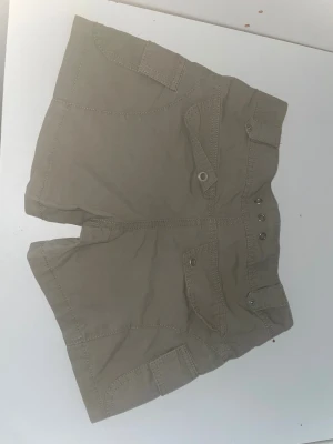 Gröna cargo shorts från Detroit - Säljer ett par beige cargo shorts från Detroit med coola fickdetaljer och knappar framtill. De har en lowaist passform och är gjorda i 100% bomull, vilket gör dem sköna och lätta. Perfekta för dig som gillar en avslappnad och streetig stil.