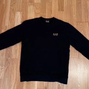 Svart EA7 sweatshirt med guldtryck - Snygg svart sweatshirt från EA7 med diskret guldigt logotryck på bröstet. Tröjan har rund halsringning och långa ärmar, perfekt för en stilren och sportig look. Mjuk och bekväm, passar till jeans eller joggers.