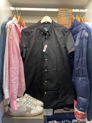 Svart skjorta Polo Ralph Lauren - Svart skjorta från Polo Ralph Lauren med korta ärmar och klassisk krage. Skjortan har knappar framtill och broderad Polo-loggan på bröstet. Perfekt stilren look till finare tillfällen. Nypris ligger runt 1500, skjortan är aldrig använd och har pristagarna kvar.                                               Skriv vid frågor, priset är inte hugget i sten🤝