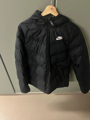 Svart pufferjacka från Nike  - Svart pufferjacka från Nike i barnstorlek 158-170, med huva och dragkedja framtill. Jackan har ett stilrent Nike-logotryck på bröstet och är tillverkad i syntetmaterial. Passformen är normal och jackan är perfekt för kalla dagar.