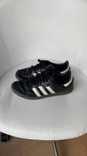 Adidas Samba svarta sneakers strl 38 - Adidas Samba sneakers i svart skinn med vita detaljer och klassiska tre ränder på sidorna. Platt sula, rund tå och snörning framtill. Ikonisk modell med retrovibe, perfekt för dig som gillar streetstyle och sportig look.