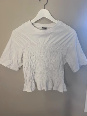 Vit smockad topp från Gina Tricot - Säljer en vit topp från Gina Tricot i storlek S. Använt en gång. Toppen har smockad midja och volangkant nedtill, samt korta ärmar och rund halsringning. Materialet är mjuk bomull och modellen är croppad med en feminin touch. Perfekt för dig som gillar stilrena och söta detaljer. 