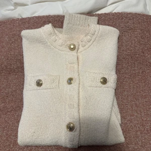 Vit kofta med guldknappar - Säljer en vit kofta med snygga guldknappar framtill och på fickorna. Koftan har rund halsringning, långa ärmar och är stickad i ett mjukt material. Perfekt för dig som gillar klassisk och clean stil med en touch av lyx.