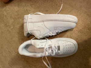 Nike Air Force 1 helvita sneakers - Nike Air Force 1 sneakers i helvit färg med klassisk design. Skorna har snörning, perforerad tå för ventilation och en platt, robust sula med cirkulärt mönster undertill. Tillverkade i skinn med mjuk vadderad insida och tydlig Nike Air-logga på hälen.