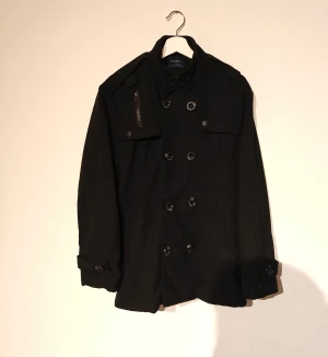 AOWOFS kappa - Svart skepparkavaj från AOWOFS med dubbelknäppning, axelklaffar och snygga detaljer som fickor med lock och en liten dragkedja på bröstet. Jackan är tillverkad i 100% polyester och har långa ärmar med knappar vid mudden. Perfekt för dig som gillar klassisk stil med en modern twist.