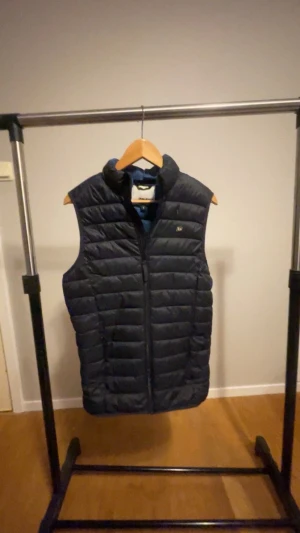 Mörkblå väst från Blend - Snygg mörkblå dunväst från Blend i storlek S. Västen har quiltad design, hög krage och dragkedja framtill. Perfekt för lager-på-lager och har en stilren look med Blend-logga på bröstet. Materialet är polyester och dun, vilket gör den både lätt och varm.