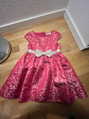 Rosa festklänning med guldrosett H&M - Superfin rosa klänning från H&M med glittrigt blommönster i guld och en stor glittrig rosett i midjan. Klänningen har kort ärm, rund hals och dragkedja bak. Perfekt för speciella tillfällen när du vill sticka ut lite extra!