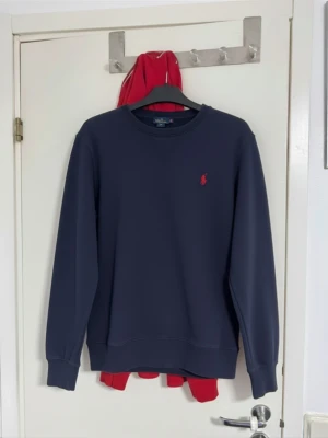 Mörkblå sweatshirt från Polo Ralph Lauren - Säljer en stilren mörkblå sweatshirt från Polo Ralph Lauren med klassisk röd logga broderad på bröstet. Tröjan har rund halsringning och långa ärmar, perfekt för en clean och avslappnad look.