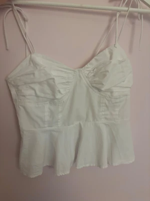 Vit linnetopp med volangkant - Säljer en söt vit linnetopp med tunna axelband som knyts och en volangkant nedtill. Toppen har lätt rynkade detaljer vid bysten och en feminin, figurnära passform. Perfekt för varma dagar och att styla med jeans eller kjol.