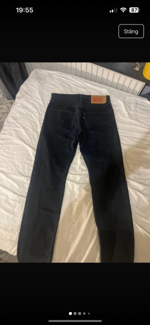 Levi's 501 svarta jeans W31 L32 - Svarta Levi's 501 jeans med klassisk rak passform och fem fickor. Jeansen har en mörk ton, tydlig Levi's patch bak och röd etikett vid bakfickan. Tillverkade i slitstarkt denimtyg av bomull. Perfekt för dig som gillar tidlös stil.