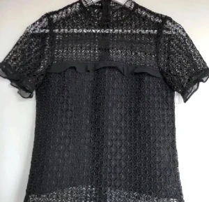 Svart spetsblus med volangdetalj - Säljer en svart blus i spets med korta ärmar. Blusen har ett genombrutet mönster och en snygg volangdetalj framtill. Perfekt för dig som gillar detaljer och vill ha en unik topp med transparenta partier.