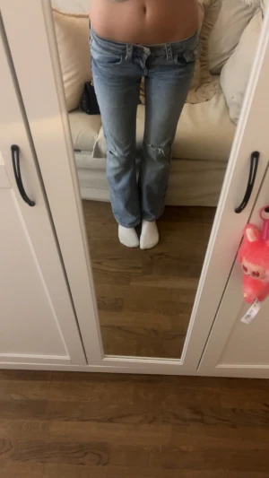 Blå bootcut jeans med slitningar - Säljer ett par ljusblå bootcut jeans med slitna detaljer på knäna och snygg tvätt. Jeansen har klassiska fem fickor, knappstängning och dekorativa sömmar på bakfickorna. Perfekta för dig som gillar en avslappnad och trendig look.