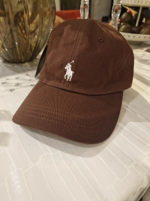 Brun keps från Polo Ralph Lauren - Snygg brun keps från Polo Ralph Lauren med klassisk vit broderad logga framtill och texten POLO baktill. Justerbar rem med metallspänne för perfekt passform. Tillverkad i mjuk bomull, perfekt för en chill och stilren look.