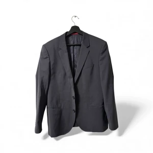 Svart kavaj från Hugo Boss - Stilren svart kavaj från Hugo Boss med klassisk passform och två knappar framtill. Kavajen har slät yta, diskreta fickor och helfodrad insida i svart. Perfekt för dig som vill ha en snygg och tidlös look. Märke syns på röd etikett i nacken.