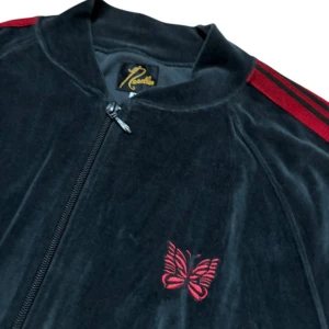 Needles svart velour track jacket - Svart track jacket från Needles i mjuk velour med röda ränder längs ärmarna och broderad röd fjäril på bröstet. Jackan har dragkedja framtill och ribbade muddar. Perfekt för en chill och sportig stil. Väldigt bra skick!