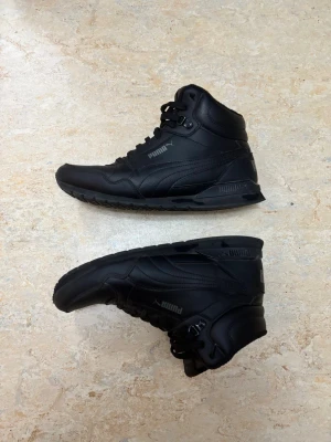 Puma ST Runner v3 Mid L - Säljer ett par svarta höga sneakers från Puma med rund tå och platt sula. Skorna har snörning och är tillverkade i skinn med mjuk vadderad insida. Puma-loggan syns på sidan och bak på hälen. Perfekta för dig som gillar sportig och clean stil.