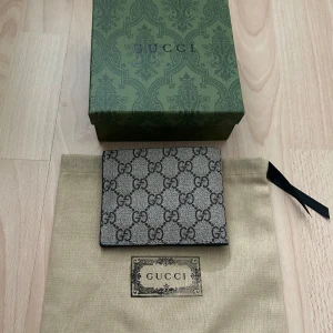 Gucci plånbok  - Snygg plånbok från Gucci i svart skinn med klassiskt GG-monogram i grått och svart. Plånboken har flera kortfack och ett fönster för ID. Kommer med original dustbag och låda, perfekt för dig som vill ha något lyxigt och stilrent i fickan. Pris kan diskuteras!!!