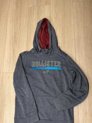Mörkgrå Hollister hoodie med vinröd huva - Snygg mörkgrå hoodie från Hollister med stor logga på bröstet och blå detalj. Insidan av huvan är vinröd och har snörning. Tröjan har lång ärm och klassisk känguruficka framtill. Perfekt för chill dagar. Mer bilder eller Frågor? Då är det bara att ge mig en signal så svarar vi oftast inom 1-2 timmar och skickas samma dag om du beställer innan 19:00 