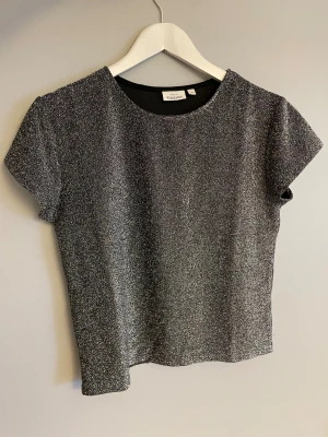 Glittrig silver topp från KappAhl - Snygg glittrig silver topp från KappAhl i storlek 170, men passar S också. Toppen har rund halsringning och korta ärmar. Materialet är mjukt och stretchigt syntet, vilket gör den bekväm att bära hela kvällen. Använd ett fåtal gånger