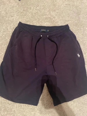 Mörkblå shorts från Polo Ralph Lauren - Snygga mörkblå shorts från Polo Ralph Lauren med klassisk broderad logga i vitt på benet. Modellen har elastisk midja med snörning och är tillverkad i mjuk bomull. Perfekta för chill dagar och har en loose passform.