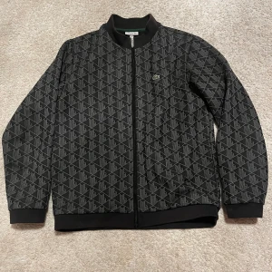 Svart bomberjacka från Lacoste, stl 14 - Snygg svart bomberjacka från Lacoste med hel dragkedja och diskret mönster över hela jackan. Klassisk ribbad krage, muddar och nederkant. Liten broderad Lacoste-logga på bröstet. Perfekt för dig som vill ha en stilren och sportig look.