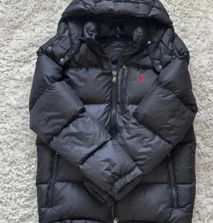 Svart dunjacka från Polo Ralph Lauren - Svart dunjacka från Polo Ralph Lauren med röd broderad logga på bröstet. Jackan har huva, flera dragkedjefickor och quiltad design. Materialet är polyester och dun, perfekt för kalla dagar. Snygg och stilren modell med klassiska detaljer.