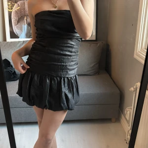 Svart axelbandslös kort klänning - svart axelbandslös kort klänning med rynkad överdel och voluminös kjol. Materialet är lätt glansigt perfekt nu till nyår eller alla julbord 🎉 använd endast en gång 