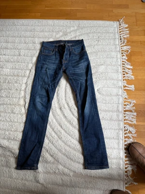 Mörkblå jeans från Nudie Jeans, 29/34 Grimtim (2008) - Säljer ett par mörkblå jeans från Nudie Jeans i storlek 29/34. Klassisk femficksmodell med kontrastsömmar och Nudie-logga på bakfickan. Jeansen har rak passform och är tillverkade i slitstarkt denimtyg.
