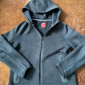 Svart hoodiejacka från Nike - Säljer en svart hoodiejacka från Nike med dragkedja framtill och två fickor med snygga detaljer. Jackan har en enkel, sportig look och är tillverkad i mjukt material som passar perfekt till chill eller träning.