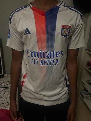 Olympique Lyonnais hemmatröja Adidas - Säljer en officiell Olympique Lyonnais hemmatröja från Adidas. Tröjan är vit med vertikala röda och blå ränder framtill, klubbmärke på bröstet och sponsortryck. Klassisk V-ringning, korta ärmar och tillverkad i lätt funktionsmaterial.