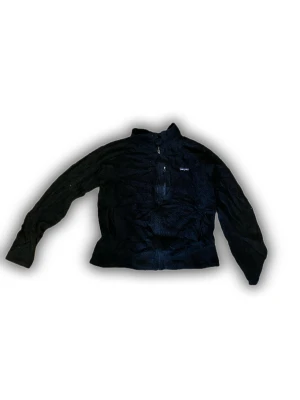 Svart teddyjacka från Patagonia - Mjuk och fluffig svart teddyjacka från Patagonia med hel dragkedja och en praktisk bröstficka med zip. Jackan har hög krage och långärmad passform, perfekt för kyliga dagar. Snyggt broderad logga på bröstet och stilren design.