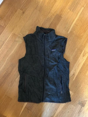 Mörkgrå dunväst från Patagonia - Snygg mörkgrå dunväst från Patagonia med hög krage och hel dragkedja framtill. Västen har två sidofickor med dragkedja och en bröstficka med dragkedja. Perfekt för lager-på-lager och outdoor. Diskret mönster i tyget och klassisk Patagonia-logga på bröstet.