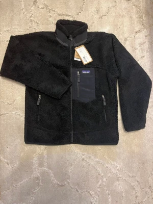 Patagonia fleece - Säljer en svart fleece i storlek M. Den är mjuk, varm och perfekt som mellanlager nu när det börjar bli kallare. Jag fick den i födelsedagspresent men den kommer tyvärr inte till användning, så jag vill gärna låta någon annan få glädje av den. Säljer den billigt och till ett riktigt bra pris eftersom jag bara vill bli av med den snabbt. Skicket är helt med tags på och aldrig använd.Hör av dig om du vill köpa eller behöver fler bilder!