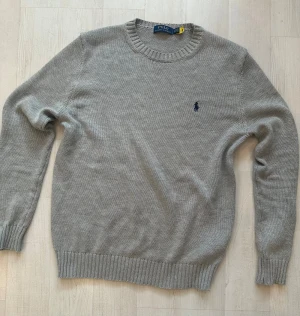 Grå stickad tröja från Polo Ralph Lauren - Snygg grå stickad tröja från Polo Ralph Lauren med klassisk rund hals och mörkblå broderad logga på bröstet. 